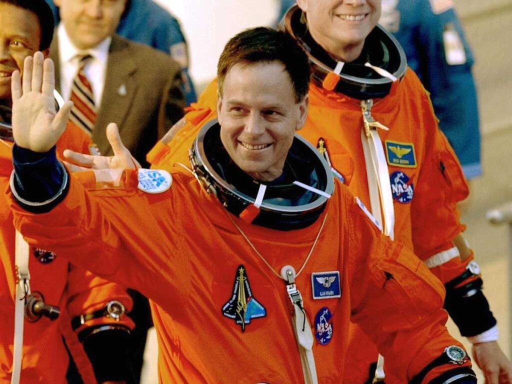 Image of Ilan Ramon in his orange NASA flight suit walking with other astronauts in the background.